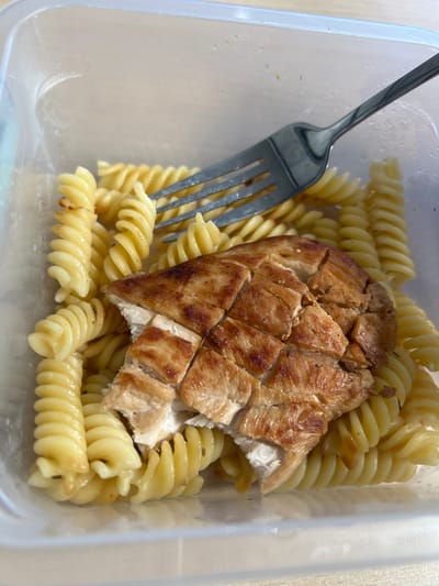 A close-up overhead shot shows a transparent plastic food container filled with fusilli pasta and sliced grilled chicken breast. A silver fork is placed on top of the food, with its tines partially submerged in the pasta. The chicken is cooked to a golden brown and appears moist. The pasta is a light yellow and has a slightly glossy sheen, indicating it may be coated in a sauce. The background is blurred, but appears to be a light-colored surface, possibly a table. There is no indication of a specific location in Sourbrodt, Belgium.