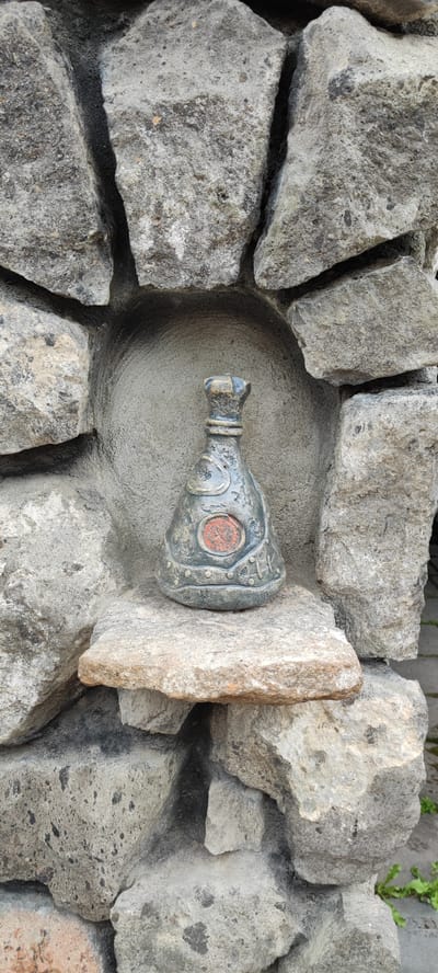 An artistic ceramic object, resembling a stylized bottle or urn with a decorative red emblem, is placed on a flat stone shelf. This shelf is recessed within a rough stone wall, forming a niche. The stone wall is constructed from irregular, grey, and light brown rocks, with visible mortar in between. The object itself has a dark, aged patina with lighter decorative elements, including a crown-like top and studded bands around its base. The background within the niche is a textured grey cement surface. To the right of the stone wall, a glimpse of green grass and grey pavement is visible. The scene appears to be an outdoor setting in Voskevaz, Armenia, likely part of a garden or architectural feature.