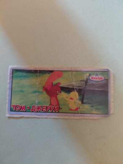 The media file contains a trading card with an illustration of cartoon characters from "Tom and Jerry." The card features Tom, a cat, and Jerry, a mouse, in a dynamic pose. Tom is depicted in mid-air, seemingly falling or jumping, with his arms and legs spread out. Jerry is not clearly visible in the immediate vicinity of Tom in this pose, but the title of the card suggests his presence. The background of the illustration is a vibrant green, suggesting an outdoor setting, possibly a garden or park. There is a hint of a dark, vertical structure in the background, which could be a tree trunk or a fence post. Above the illustration, a circular logo with the text "Henry" in a stylized font is visible, indicating a brand or series. Along the right edge of the card, text in a colorful, stylized font reads "ТОМ И ДЖЕРРИ," which is the Russian transliteration of "Tom and Jerry." The card itself is rectangular with rounded corners and appears to be made of thick paper or cardboard. It has a somewhat worn appearance, with visible creases and minor damage to the edges, suggesting it has been handled or used. The overall impression is that of a collectible item, likely from a children's cartoon series. The light source seems to be from above and slightly to the left, casting subtle shadows and highlighting the colors of the illustration. No specific time of day or weather conditions are evident. The context of Marynivka, Ukraine, provides a geographical location for the origin or use of this item, but no direct visual cues within the image itself point to this specific location.