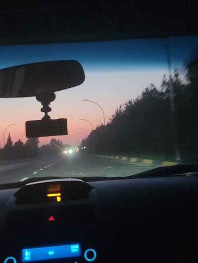 The image captures a view from inside a car driving on a road at dusk. The sky exhibits a gradient of colors, transitioning from a deep blue at the top to soft pinks and oranges near the horizon, suggesting sunset or sunrise. Silhouetted trees line the right side of the road, with their forms blurred due to motion. Streetlights with curved tops are visible along the roadside. The road itself has white dashed lines marking the lanes, and on the right, there are yellow and white markers delineating the edge. In the distance, headlights from oncoming vehicles are visible. Inside the car, the dashboard is partially visible. A rearview mirror is mounted at the top, with a black dashcam attached below it. An orange digital display is visible on the dashboard, showing what appears to be a directional arrow and a number. A red triangular warning light is also illuminated on the dashboard. Below this, a blue display shows icons and some text, including what might be time and temperature. The overall scene depicts a journey through a semi-urban or rural setting during twilight. The location is identified as Kipchak, Uzbekistan.
