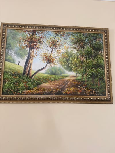 This image presents a framed oil painting, photographed at a slight downward angle against a plain, light-colored wall. The painting, horizontally oriented, is enclosed within a decorative gold-colored frame, characterized by an ornate, repetitive beaded pattern along its borders.

The painting itself depicts an autumnal forest landscape. A dirt path, featuring two distinct parallel tire tracks, recedes diagonally into the background from the lower center, covered in a dense scattering of fallen leaves in shades of yellow, orange, and brown. To the left of the path, the terrain rises gently, covered in vibrant green undergrowth interspersed with additional fallen leaves.

Two prominent trees are positioned on the left side of the path in the midground. The leftmost tree possesses a robust, dark brown trunk and branches adorned with rich reddish-orange autumn foliage. Adjacent to it, a second tree with a more slender, lighter-colored trunk curves gracefully towards the path, also displaying reddish-orange and yellow leaves. On the right side of the path, a dense succession of trees with various green and yellowish-green leaves forms a natural corridor, extending into the background. The background is characterized by a soft, hazy blue light, indicative of distance or mist, with the silhouettes of additional trees. A clear, light blue sky is visible above the tree line, particularly towards the upper left.

A signature, rendered in a non-Latin script followed by characters resembling "02" or "04", is visible in the lower right corner of the painting.

This image, which portrays the aforementioned painting, is located in the city of Aragats, Armenia. No individuals or specific actions are depicted within the painting. The immediate surrounding environment visible in the photograph is a featureless, light-toned wall.