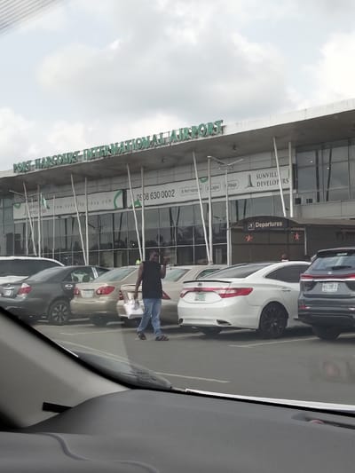The image depicts the exterior of Port Harcourt International Airport in Port Harcourt, Nigeria, as viewed from inside a vehicle. The airport terminal building dominates the background, featuring a modern design with a prominent "PORT HARCOURT INTERNATIONAL AIRPORT" sign in green letters above a facade of dark, reflective glass windows. The building's overhang is supported by multiple slender, white, tapered pillars. Additional signage for "SUNVILLE DEVELOPERS" and a "Departures" sign are visible.
In the midground, a paved parking lot contains several parked passenger vehicles, including a dark grey sedan, a gold/beige sedan, a white Hyundai Azera/Grandeur with black rims, and the rear of a dark grey SUV with the license plate "MUG 195 JS" partially visible.
A single male individual is walking across the parking lot from left to right. He is of dark complexion, wearing a dark sleeveless shirt, blue jeans, and dark sandals. He carries a white bag in his left hand and has his right hand raised, appearing to gesture or hold an object, while looking towards the viewer. The sky is overcast with uniform grey clouds. The foreground shows a portion of a vehicle's dashboard and windshield, indicating the vantage point of the photographer.