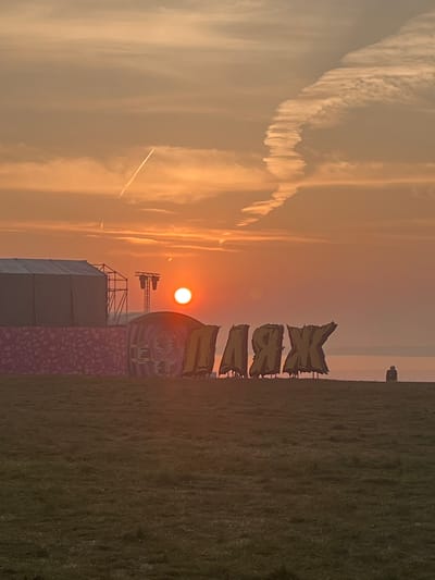 The image shows a sunset over a grassy field with a stage structure in the background. The sun is a bright orange disc low on the horizon. There are large, yellow letters spelling out "KRAX" in front of the stage. A single person is sitting on the horizon to the right. The sky has wispy clouds and contrails from an airplane.