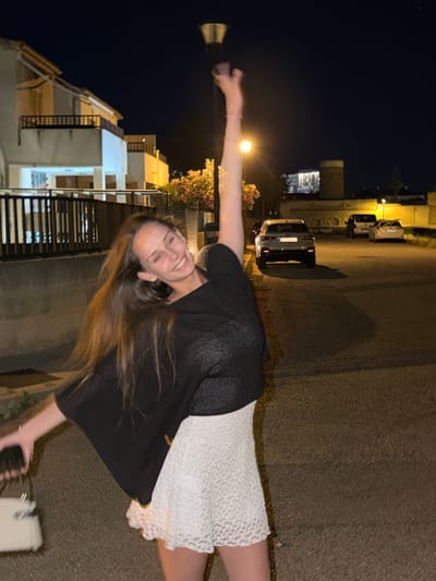 The image shows a young woman standing on a street in Platja D'alcúdia, Spain. She is wearing a white dress and a black shawl. She is holding a phone in one hand and sunglasses in the other. In the background, there are buildings, cars, and streetlights. The time of day appears to be night.