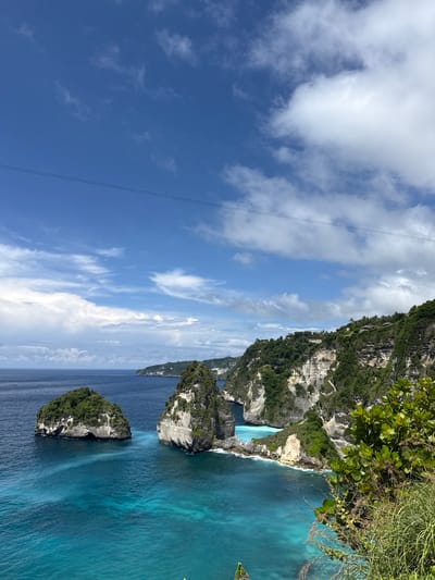 The image captures a scenic view of Kelingking Beach, a well-known landmark located in Pejukutan, Indonesia. The camera angle is tilted, showcasing a blend of natural elements.
The landscape features a towering cliff covered in lush greenery, contrasting sharply with the light-colored, tiered rock formations along the shore. The cliff drops off into a turquoise body of water, clear and inviting. In the foreground, smaller islands, also covered in vegetation, are surrounded by the ocean. The sky is a brilliant blue, dotted with white, fluffy clouds. Overhead electrical wires are visible, cutting across the sky.