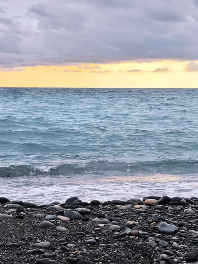 This is a photograph of a beach with a pebble shore and the sea. The sky is overcast with a band of yellow light at the horizon, suggesting either sunrise or sunset. The sea is a dark blue with small waves lapping at the shore. The shore is covered in dark grey pebbles and larger smooth stones. There are no people or animals visible in the image. The scene depicts a natural coastal environment. The location is Zemo Kvariati, Georgia.