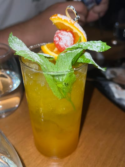 A close-up, eye-level shot shows a tall glass of amber-colored liquid, likely a cocktail, filled with ice. The drink is garnished with a skewer of two orange slices, one dusted with powdered sugar, and a red berry, also dusted with sugar. Several sprigs of fresh mint, dusted with powdered sugar, are also arranged around the rim of the glass. The drink is placed on a wooden surface, likely a table. In the background, out of focus, a person's arm is visible. The overall lighting is dim, suggesting an indoor setting, possibly a restaurant or bar in Chaidari, Greece.