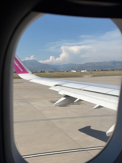 A view from an airplane window shows a white airplane wing with a pink stripe on its edge, bearing the text "wizzair.com." The wing is positioned over a concrete tarmac with yellow and white markings, typical of an airport runway or taxiway. In the background, a grassy field transitions to rolling hills and mountains under a bright blue sky with scattered white clouds. The airport appears to be located in a semi-arid environment with sparse vegetation. There are no people visible. The image captures a moment of preparation for takeoff or after landing at an airport near Donja Cijevna, Montenegro, evidenced by the distinct mountain range in the distance and the overall landscape.