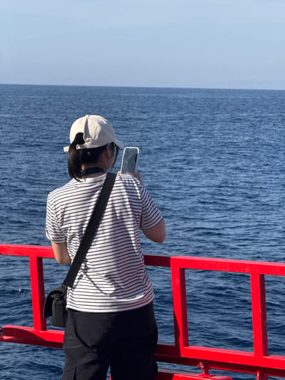 A person wearing a white baseball cap, a black and white striped t-shirt, and black pants stands on a red railing, holding a silver smartphone to take a photo of the ocean. The person has a black crossbody bag slung over their shoulder. The ocean is a deep blue with gentle waves. The sky is a clear, pale blue. The location is the city of Dong Son, Vietnam.