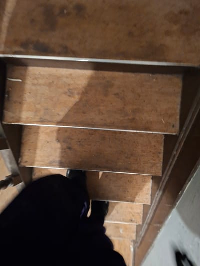This is a close-up, downward-angle shot of a person descending wooden stairs. The person is wearing dark clothing, including a dark purple or blue garment visible on their left shoulder and dark shoes. The stairs are made of a light-colored wood with a visible grain and appear somewhat worn. Each step has a thin metal or silver strip along its edge. The lighting suggests an indoor environment, possibly a basement or attic, with the light source appearing to be from above or to the side, casting shadows on the stairs. The overall impression is functional rather than decorative, with a focus on the act of movement down the staircase. No visible text is present. The location context is Evere, Belgium.