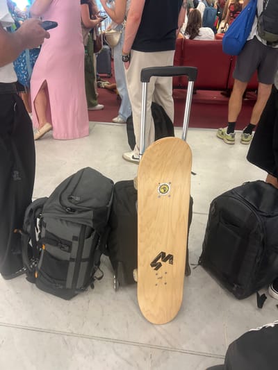 The image depicts a floor scene, likely within an airport terminal, based on the presence of luggage and the tiled floor. In the foreground, a light-colored wooden skateboard with black graphics is propped upright, secured to a rolling suitcase. To the left of the skateboard, a large gray and black backpack rests on the floor, with another black backpack situated to its right. The feet of several individuals are visible. A person on the left wears dark pants, and another person to the right wears dark shorts and lime green athletic shoes with black socks. Further back, a person in a long pink dress is partially visible, as are other individuals and luggage, including a blue duffel bag and red seating. The environment suggests a public waiting area. This scene could be located in Paray-Vieille-Poste, France, if this is a public space within the jurisdiction of that city, such as a transportation hub.