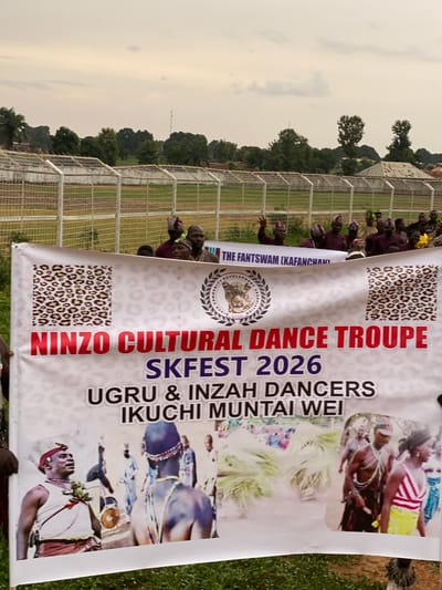 The image captures a banner displaying the text "NINZO CULTURAL DANCE TROUPE SKFEST 2026 UGRU & INZAH DANCERS IKUCHI MUNTAI WEI." The banner also includes images of people performing cultural dances.
A group of people stands behind the banner, some wearing traditional headwear. A chain-link fence stretches in the background. Trees and a cloudy sky are visible above the fence. The banner is placed in the city of Kafanchan, Nigeria. The text on the banner also contains the words "THE FANTSWAM (KAFANCHAN)."