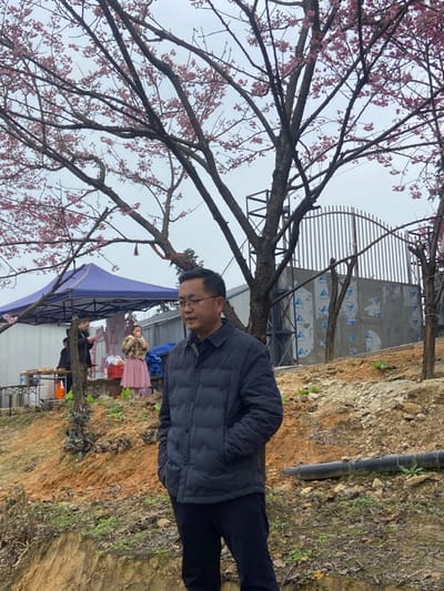 An Asian man with glasses and a dark jacket stands outdoors. He faces left, beside a tree with pink blossoms. In the background, a blue tent and people are visible. The location seems to be a garden. A hill slopes upward to the right side of the image, where a black pipe runs down the surface. The sky is overcast.