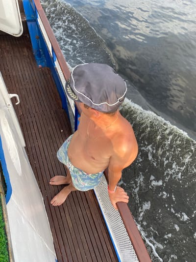 A man wearing a captain's hat and swim trunks stands on a wooden deck, gripping a railing. He is looking down at the water as a boat speeds along, creating a wake. The sun is shining, and the weather appears to be warm. The man has a golden chain around his neck and a pink band on his wrist. The boat is blue and white, with a wooden deck. The water is dark and choppy. The scene appears to be set on a sunny day in Russia.