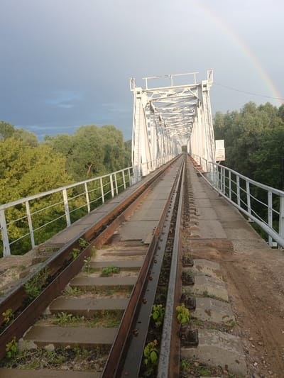 This image shows a railway bridge in Pakaliubičy Rural Council, Belarus. The bridge is made of white metal and has two sets of railway tracks. The tracks are made of steel and are laid on concrete sleepers. The bridge is surrounded by trees and greenery. In the background, a rainbow is visible in the sky. The sky is partly cloudy. The lighting suggests it is either early morning or late afternoon. There are no people or animals visible in the image. The scene appears to be peaceful and natural.