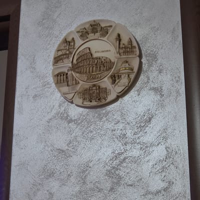 This is a close-up, eye-level shot of a decorative plate, likely a souvenir, mounted on a textured, light-colored wall. The plate is round with scalloped edges, and it is divided into sections, each featuring a bas-relief carving of a famous Roman landmark. The central section prominently displays the Colosseum, with the word "ROMA" inscribed in a circle around it. Other sections depict structures such as the Pantheon, Castel Sant'Angelo, and St. Peter's Basilica. The plate is a light, neutral color, possibly cream or beige, and the carvings are detailed, with some areas highlighted with a gold-like sheen. The wall has a rough, stucco-like texture with a shimmering quality, suggesting it might be decorative plaster with metallic accents. A dark wooden frame runs along the top of the image, partially obscuring the top of the wall. The lighting is even, and there are no people or clear indications of time of day or weather. The scene is static and focuses on the decorative object and its backdrop. The visible text on the plate reads "ROMA", "COLOSSEO", and "CASTEL SANT'ANGELO". The location context of Peshtera, Bulgaria, is noted but not directly visible in the image.