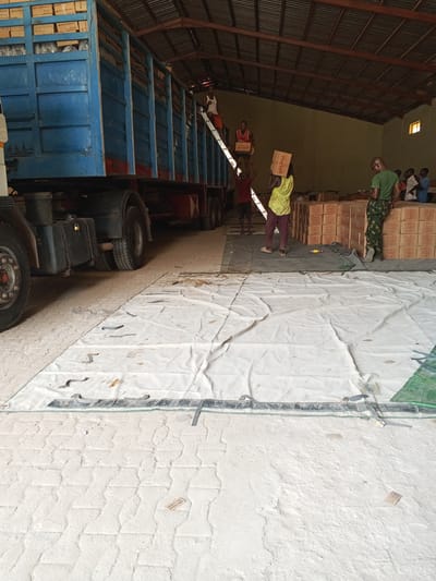 The image depicts a group of individuals unloading boxes from a blue truck inside a warehouse in Pasere, Nigeria. One man is on a ladder inside the truck, handing boxes to others on the ground. The boxes appear to be stacked in organized rows near the back of the warehouse. There are a number of boxes already unloaded. A white tarp covers a portion of the floor in the foreground. A person is visible, dressed in military clothing.