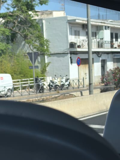 This media file captures a scene from inside a vehicle, looking out through the windshield. The image is oriented vertically, giving a somewhat skewed perspective. In the foreground, a dark, curved object, likely part of the vehicle's interior, dominates the right side of the frame.
The background reveals a street scene in what appears to be a European town, possibly Santa Eulària des Riu, Spain, as indicated by the context. Across the street, a multi-story building with white and gray facades is visible. The building has numerous windows and balconies. Some windows are open, and there are hints of interior spaces. Above the building, a vibrant patch of pink and red flowers, likely bougainvillea, adds a splash of color.
Several motorcycles are parked neatly along the sidewalk in front of the building. They are lined up and appear to be of similar models, suggesting a parking area for residents or patrons. A traffic sign with a blue circle and red diagonal line is mounted on a pole, indicating a traffic regulation. Further down the street, a white vehicle, possibly a delivery van or a small truck, is partially visible.
Green foliage from trees and shrubs is scattered throughout the scene, particularly in the lower left portion of the image, and peeking from behind the building. The sky, visible in the upper left, is a clear blue, suggesting fair weather and daylight. The overall impression is a glimpse of everyday life in a Mediterranean town, with parked motorcycles, a residential building, and lush vegetation. No people are clearly discernible in the frame.