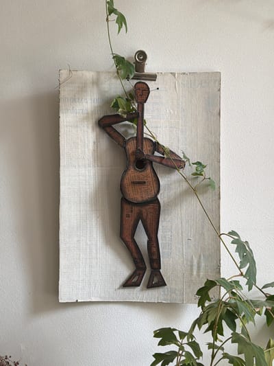 The image shows a piece of artwork hanging on a white wall. The artwork consists of a rectangular board with a textured white surface and visible writing underneath. A wooden figure in the shape of a person playing a guitar is affixed to the board. The figure appears to be made from wood or cardboard with printed text on its surface. A metal clip is attached to the left side of the board. A vine with green leaves partially covers the artwork, extending from the left side and across the top right corner. The location is not discernible from the image. Therefore, I am unable to provide information about Aubervilliers, France.