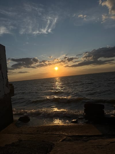 This is a photograph of a sunset over the Black Sea in Zatoka, Ukraine. The sun is low on the horizon, casting a golden light across the water and illuminating the clouds. The waves are small and gentle, lapping at the shore. In the foreground, there is a concrete structure on the left and two tires stacked on the right. The overall mood is peaceful and serene.