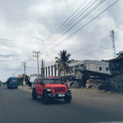 The image captures a daytime scene on a road in Payameuneng, Indonesia, under an overcast sky. The main subject is a bright red Jeep Wrangler Rubicon with the license plate "B 699 YUN", driving in the foreground towards the viewer's right. To the left, a blue box truck is also in motion on the asphalt road, heading in the same direction.
The setting is an urban or semi-urban street lined with buildings and various infrastructure. Overhead, numerous power lines crisscross the sky, supported by multiple utility poles visible along the road. A tall cellular or telecommunication tower stands prominently on the right side of the frame, piercing through the cloudy sky.
On the right side of the road, there's a multi-story white building, possibly a commercial establishment or office. In front of it, a lush green palm tree stands tall. Adjacent to the white building are smaller, more rustic structures that appear to be shops or workshops, with various items like stacked curved frames and construction materials visible around them. A large white banner displays "DETAYA" and "coffeeshop". Nearby, a poster with images of people in traditional attire is also visible.
Further down the road on the left, a yellow sign with "Alfamart" and "DOPS" (likely "SUPERMARKET" below Alfamart) indicates a convenience store. The general activity is daily traffic on a cloudy day in a populated area.