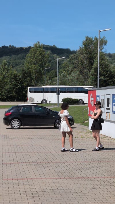 The image depicts an outdoor scene with two young women standing near a wall. The woman on the left wears a white dress with puff sleeves and a floral pattern, white socks, and black sandals. She has curly dark hair and is looking away from the camera. The woman on the right is wearing a black skirt, a multi-colored patterned top, white socks, and black sandals. She has dark hair and is looking towards the woman on the left, with her arms crossed. They are standing on a paved area with interlocking bricks.
In the background, a black hatchback car is parked to the left, and a white bus is parked further back. Behind the bus, there is a line of trees and a hilly landscape under a clear blue sky. The wall behind the women features several signs, including one with a red "etu" logo and other symbols, possibly indicating services. This location appears to be a rest stop or a parking area near a road. The overall environment suggests a sunny day in a semi-rural or roadside setting. No specific landmarks in Cornu de Jos, Romania, are identifiable from the image.