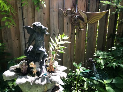 A garden scene depicts several decorative figurines and ornaments against a weathered wooden fence. On the left, a large, dark-colored statue of a winged figure, possibly an angel or a fairy, sits on a textured, light-colored stone fountain. At the base of this statue, nestled in the fountain, are several smaller figurines, including two fairies in pink and orange dresses and a tiny blue and white figure. To the right of the fountain, a metallic, gold-toned mask with swirling accents is mounted on the fence, featuring a face with stylized hair and wings. Further to the right, a dark-colored figurine of a person or creature with textured hair and clothing is perched on a shiny green, leaf-shaped object. The background consists of lush green foliage and the vertical wooden planks of a fence. The scene is illuminated by natural sunlight, casting shadows. The location is identified as the city of Severn, Canada.
