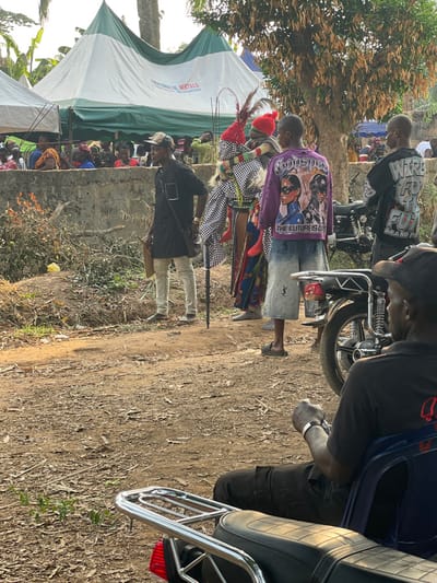 The scene depicts an outdoor community gathering in Umueze, Nigeria, under daylight conditions.
In the immediate foreground, a dark-skinned man is seated on a blue plastic chair, viewed from behind and to the left, wearing a black short-sleeved shirt and dark pants. A silver bracelet is visible on his left wrist. The rear section of a black motorcycle, including a chrome luggage rack and taillight, is positioned in front of him. The ground is unpaved dirt, sparsely covered with dry leaves and some vegetation.
The midground features several individuals. From left to right: A man stands facing slightly right, wearing a black long-sleeved tunic, light-colored pants, a cap, and eyeglasses. He carries a dark cross-body bag and holds a dark, slender staff in his right hand and a light-colored object in his left. Behind him is a concrete wall. Immediately to his right stands a masquerade figure dressed in a black-and-white checkered garment over a multi-colored patterned skirt. Its headpiece is red with prominent feathers, and small red fabric elements dangle from its costume. To the right of the masquerade, a man is seen from behind, wearing a purple long-sleeved shirt featuring graphic prints of three figures and partially visible text including "THE FUTURE IS..." and "GOOSPEED." He wears light-colored, knee-length patterned shorts and dark sandals. Further right, another man is viewed from behind, wearing a black t-shirt with white text that reads "WIRE FOR THE FUTURE BRAND." Partial views of motorcycles are present among these individuals.
In the background, behind the concrete wall, a large white and green canopy tent is visible, bearing the branding "PRAGMACH RENTALS" and contact information. A crowd of people, many dressed in colorful clothing including red, are gathered beneath and around this tent. Trees with sparse foliage are visible throughout the background.