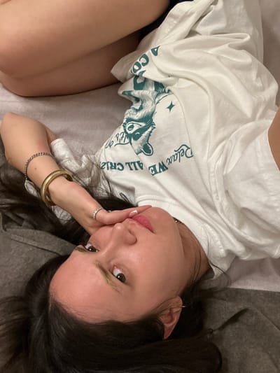 The image displays a single female individual, viewed from an elevated perspective, reclining on a light-colored soft surface indoors. Her upper torso and head are visible, with her dark hair splayed against the light gray fabric. She has a fair complexion, and her head is turned towards the camera, eyes open. Her right hand is positioned near her mouth, with fingers slightly curled; a silver-toned ring with multiple small stones is visible on her right ring finger. Her left wrist is adorned with a silver-toned chain bracelet and a wide, gold-toned ribbed bangle. She wears a white short-sleeved t-shirt featuring a green graphic print of an animal's head (possibly a raccoon) and partial text that includes "Relax WE" and "ALL CR". A dark fabric, likely part of her lower garment, is partially visible beneath her legs. The environment consists solely of the soft, muted gray fabric she is resting on, with a darker gray section near her upper left. No specific architectural elements or landmarks are present to confirm the location in Vandœuvres, Switzerland.