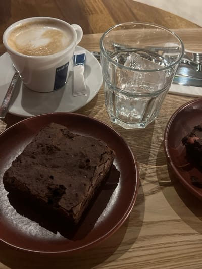 The image displays a tabletop scene within an establishment located in Lordelo do Ouro e Massarelos, Portugal. The surface is a light-colored wooden table. A white ceramic cup bearing a blue "Lavazza" logo contains a cappuccino with distinct foam art and rests on a white saucer. A silver spoon and a "Lavazza" branded sugar packet are positioned on the saucer. Adjacent to this, a clear, faceted glass tumbler holds water and several ice cubes. In the lower left, a dark chocolate brownie with a cracked surface is presented on a dark red circular plate. A second dark red plate, partially visible in the lower right, contains a partially consumed chocolate brownie, which exhibits a reddish inclusion. Objects in the background, including cutlery and a white napkin, are out of focus. No individuals are present in the frame.