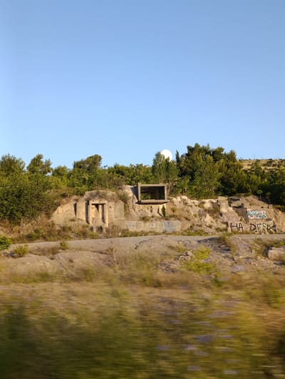 The image shows a clear, bright day with a pale blue sky. In the foreground, the landscape is blurred due to motion, appearing as streaks of green and brown vegetation. Behind the blurred foreground, a rocky hillside is visible.
On the hillside, there are remnants of concrete structures, some of which are covered in graffiti. One structure is an open-sided rectangular frame, and another is a smaller, more enclosed structure with an entrance. The concrete appears weathered and aged.
Above and behind the concrete structures, there is dense green foliage from trees and shrubs. In the distance, on the crest of the hill, a white, dome-shaped structure is visible, possibly a radar station or similar facility. The overall impression is one of an abandoned or disused site, likely in a natural or semi-natural environment. The location is identified as Donja Cijevna, Montenegro.