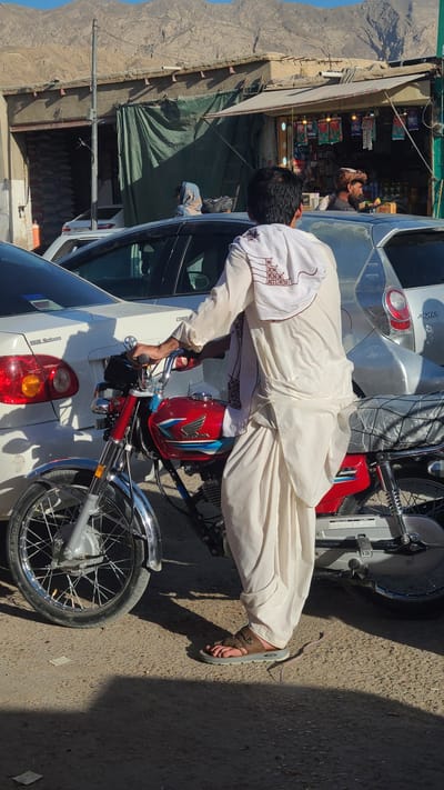 A man dressed in a traditional white shalwar kameez and brown sandals stands on a street next to a parked white car and a red and silver motorcycle, possibly making repairs or adjustments to the motorcycle. The street scene includes a storefront with merchandise displayed inside, and in the background, there is a mountainous landscape. The location is identified as Kuchlak, Pakistan, based on context, and the image is shot from a slightly elevated angle, possibly from the opposite side of the street.
