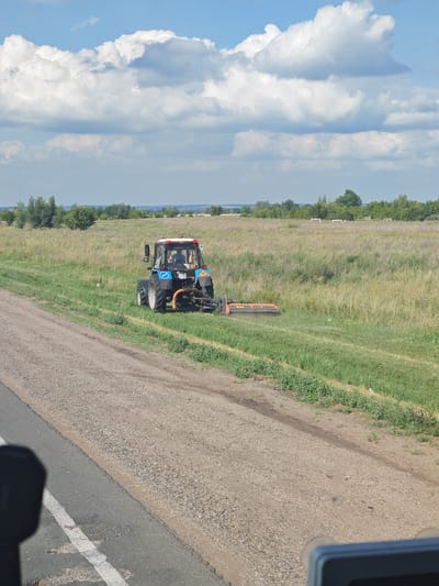 A blue tractor with a red light on top is mowing a field of green grass beside a dirt road. The tractor has a person inside, visible through the glass cab. The tractor is equipped with an orange mower attachment. The scene is set in a rural, open field with trees and vegetation in the background. The sky is blue with large, white clouds. The time of day appears to be daytime, likely mid-morning to afternoon, based on the lighting and clear sky. The weather seems to be fair and sunny. The location is identified as Chernovskii, Russia. The image captures an agricultural activity, likely for farming or maintenance of the land. There is visible text on the tractor's license plate which reads "TN 67 47".