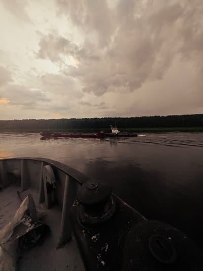 The image depicts a wide river scene at dusk, with a dramatic, cloudy sky overhead. In the foreground, the deck of a vessel is visible, with metal railings and some debris. Further out on the water, a red barge is being pushed by a tugboat. The barge is positioned on the left side of the tugboat, and both vessels are leaving a wake on the dark water. The shoreline on the opposite side of the river is lined with a dense, dark green forest. The lighting suggests either sunrise or sunset, with diffused light filtering through the clouds. The location is identified as the city of Kazachinskoye, Russia.