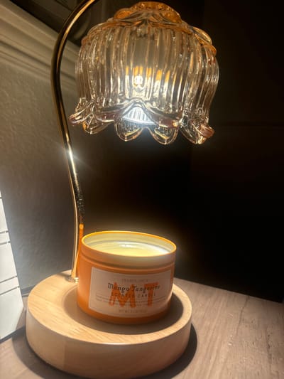 The image displays a lit electric candle warmer lamp illuminating an unlit candle, set indoors in Rancho Murieta, United States. The lamp features a golden metallic curved arm supporting a clear, ribbed glass shade with a floral motif, from which an illuminated bulb emits a warm light downwards. Directly beneath the lamp, an orange metallic tin contains a "Trader Joe's Mango Tangerine Scented Candle," with solid, yellow-cream wax. This candle rests on a light-toned, circular wooden base that also anchors the lamp's structure. The immediate background to the left reveals a partial white tiled surface, while the right and rear backgrounds are dark and featureless. The entire arrangement is situated on a darker, textured surface. No people are visible.