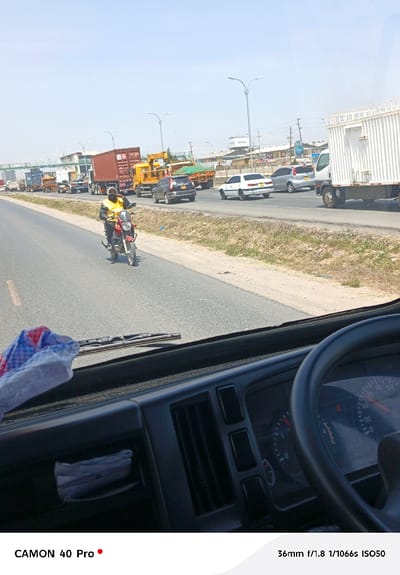 A traffic jam is seen from inside a vehicle, with a motorcyclist in a yellow vest riding on the left side of the road. Numerous trucks and cars are lined up ahead, indicating a slowdown. The driver's-side dashboard and steering wheel are visible in the foreground, adding perspective. Sunlight streams through the windshield, contributing to the scene's daytime ambience. A building in the background is partially obscured by the traffic.