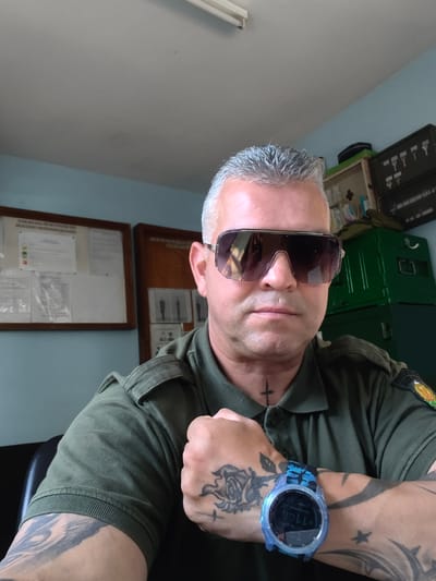 The image is a medium shot, selfie-style portrait of a man in a green collared shirt. He is wearing large, reflective sunglasses and has short, grey hair. A small, black cross tattoo is visible on his neck. His left arm is bent, and his fist is clenched. His forearm and bicep are covered in tattoos, including a depiction of a rose. A blue and black digital watch is worn on his left wrist.
The man is seated in an indoor setting that appears to be an office or administrative room. Behind him, to the left, are two bulletin boards with documents and some framed images. To the right, there is a light blue wall, and further back, a dark green metal cabinet with several compartments. A first aid kit is visible on top of a lighter green cabinet to the left of the metal one. The overall lighting suggests an indoor space with natural light coming from the front and artificial lighting from above. The image does not provide specific details that would allow for definitive location identification within the city of Bozhurishte, Bulgaria, beyond the general context of an office or similar establishment.