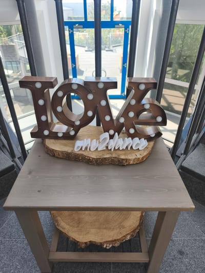 A rustic wooden table stands in what appears to be a modern building with large windows, possibly a public space or a reception area. The table is topped with a thick, round slice of wood, on which a large, illuminated metal sign spelling "LOVE" is placed. The sign is a dark metallic color with small, white, bulb-like lights embedded in it. In front of the "LOVE" sign, a white 3D text reads "one2dance." In the background, through the windows, glimpses of greenery and architectural elements of a city can be seen, including a blue-framed doorway and railings. The overall setting suggests a decorative display for an event or venue in Niederkassel, Germany.
