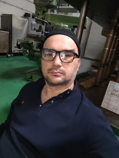 A man wearing a black beanie and glasses is pictured in a well-lit industrial setting. He is looking directly at the camera, with a slight smile. The man is wearing a dark blue polo shirt. In the background, a large green industrial machine is visible, along with metal pipes and a white sheet of paper with writing on it. The floor is green. The overall impression is of a workshop or factory environment.