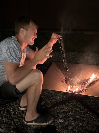 A man, wearing a patterned t-shirt and dark shorts, is sitting on a chair next to a fire pit at night. He is holding a stick over the fire, which is burning wood and embers. The man is smiling and appears to be engaged in an activity with the fire. The surroundings are dark, with gravel on the ground. The image appears to be taken in the city of Starokorsunskaya, Russia.