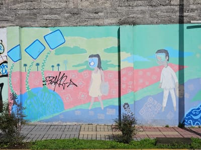 The image is a photograph of a mural on a wall in Osorno, Chile. The mural is divided into two sections. The left section depicts a figure with a blue square head and a "thumbs up" icon on its face. The figure is wearing a light-colored dress and carrying a bag. The background of this section features blue square shapes resembling screens with palm trees. The right section depicts a figure with a similar blue square head filled with an image. The figure is wearing white clothing. The background includes blue cloud shapes. The bottom of the mural has a blue section with white QR codes. Graffiti covers part of the mural. The wall has a rough concrete top. Bushes grow in front of the wall, and a sidewalk with grass separates the mural from the street.