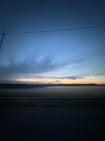 An image captured during twilight shows a vast expanse of sky transitioning from a deep blue to lighter hues near the horizon, where a faint glow of sunrise or sunset is visible. Wispy clouds are scattered across the sky, some appearing darker and more textured than others. A thin, dark horizontal line, possibly a wire or cable, stretches across the upper portion of the frame. Below the sky, a dark, silhouetted treeline forms a distinct horizon. In front of the treeline, a low-lying mist or fog blankets a landscape, obscuring finer details of the terrain. The foreground features a dark, blurred horizontal band, suggestive of a road or barrier, and below that, a deep, dark expanse that is largely indistinguishable. There are no people or animals visible in the image, and no discernible actions or interactions are taking place. The overall environment appears to be rural or semi-rural, characterized by open land and natural elements, possibly taken from a moving vehicle given the blur in the foreground. The location in Klenovskoe, Russia, suggests a natural landscape scene characteristic of the region.