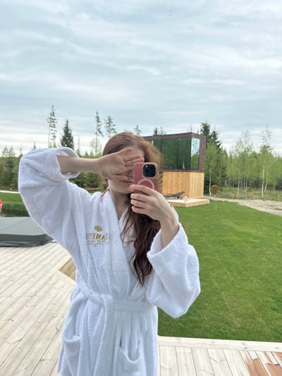 A person wearing a white bathrobe is taking a selfie outdoors. The location appears to be a wooden deck next to a lawn. Behind the lawn is a small, modern wooden structure with large glass windows. In the background are trees and a cloudy sky. The person is holding a pink smartphone in their right hand, partially obscuring their face with their left hand. The bathrobe has a logo on the chest. The scene appears to be in Vaibla, Estonia, based on the location information.