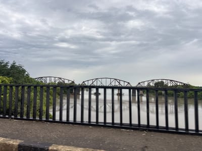 The image depicts a cloudy day with three truss bridges spanning a wide, muddy river. The bridges are made of dark metal and have a distinct arched structure. In the foreground, there is a black metal railing and a paved road surface, suggesting the photo was taken from another bridge or elevated walkway. Lush green trees line the banks of the river on both sides. The sky is overcast with a thick layer of gray clouds, casting a diffused light over the scene. No people are visible in the image. This scene likely represents the Rioni River bridges near Samtredia, Georgia, which is known for its railway junctions and bridges over the Rioni River.