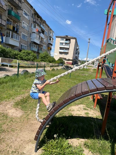 In Кульдур, Russia, on a sunny day, a young boy in a patterned bucket hat, striped t-shirt, and blue shorts is on a playground. He is seated on a brown wooden curved structure, holding onto a thick white rope. The playground equipment, with orange metal supports, is set against a backdrop of residential buildings. One building is a multi-story concrete structure with balconies, some adorned with laundry. Another building, further in the distance, appears to be made of brick. The ground is a mix of sand and green grass, with a few dandelions. The sky is bright blue with scattered white clouds.