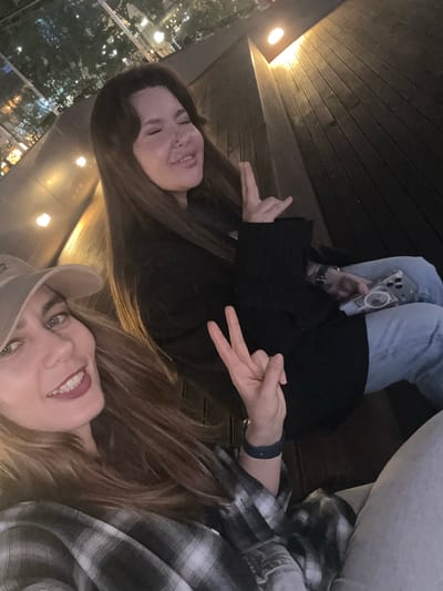 A selfie captures two women smiling and posing in an outdoor or semi-outdoor evening setting in Gimpo-si, South Korea. The perspective is tilted, showing the subjects leaning back or lying down on a surface of dark wooden planks.
In the foreground on the right, a woman with long, light brown hair wears a black and white plaid shirt and a light-colored baseball cap. She smiles broadly, revealing red lipstick, and her right hand is raised, making a peace sign. A dark wristband is visible on her left wrist.
Behind her and to the left, another woman with long, dark brown hair smiles and has a nose piercing. She wears a black jacket or top and light blue jeans. Her left hand is visible, making a gesture with her index and pinky fingers extended. She holds a smartphone with a clear case and a circular magnetic attachment.
The background shows blurred city lights and what appears to be foliage or trees through a glass barrier, suggesting an elevated viewpoint like a balcony or rooftop deck. Warm artificial lights illuminate the wooden structure, indicating it is nighttime or late evening. Both women appear relaxed and happy. No visible text is discernable in the image.