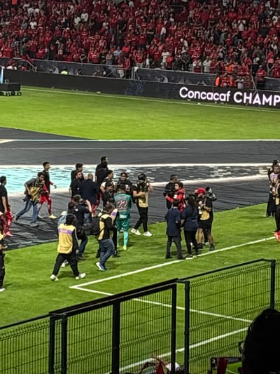 The image shows a soccer game field with the words "Concacaf Champ" vertically displayed on a sign on the left. On the far left is a large crowd of spectators, predominantly wearing red. A group of individuals, including soccer players in uniform and people wearing yellow vests labeled "Team," are gathered on the field. Some players are hugging. A black tarp is spread on the field. A green mesh fence is on the right. This is likely a game in Toluca, Mexico, based on the red team and the "Concacaf" signage.