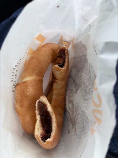 The image depicts two elongated, light brown pastries, identified as specific to the city of Costinești, Romania, held within a crumpled white paper bag. One pastry remains largely intact, exhibiting a curved shape. The second pastry is broken open, revealing a dark brown, viscous filling, suggesting a sweet paste such as chocolate. The interior of the paper bag displays partial black text, including the Romanian words "TRADIȚIE" (tradition) and "ATATEA". An orange, partial 'U'-shaped design or logo is also visible on the bag's right side. Dark blue fabric, indicative of clothing, is visible on the extreme left and right edges of the frame, suggesting a person is holding or resting the bag. No individuals are directly visible. The scene is presented from a slightly elevated, downward perspective, focusing on the contents of the bag.