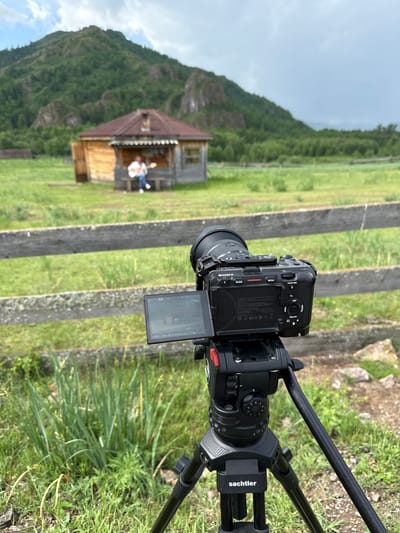 In Anchyl-Chon, Russia, a camera on a Sachtler tripod captures a scene featuring a person playing a guitar in front of a small wooden house. The house has a dark roof and wooden walls, with a porch area where the person is seated on a bench. Behind the house, a lush green mountain range covered in trees rises under a cloudy sky. A weathered wooden fence runs horizontally across the mid-ground of the image, separating the foreground with the camera from the rural landscape. The camera itself is a Sony model with a lens attached, and its screen is visible, displaying the framed shot. The ground is covered in green grass and some wild vegetation.