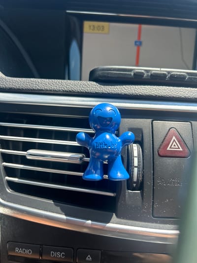 In Belokopitovo, Bulgaria, a blue, anthropomorphic air freshener with a smiley face and the text "Little.be" is clipped to a car's air vent. The air freshener is positioned on the left side of the vent. To the right of the air freshener, a red triangular hazard light button is visible. Below the air vent, car dashboard buttons are partially visible, labeled "RADIO" and "DISC". In the background, the car's navigation screen displays a map with a route indicated by a red line, and the time "13:03". A smartphone is resting on the dashboard above the navigation screen. The car's interior appears to be in daylight.